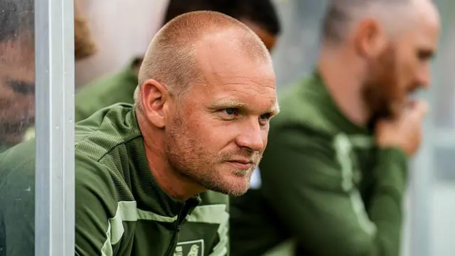 Liam Manning stares ahead while sat in a dugout wearing a green Norwich training kit