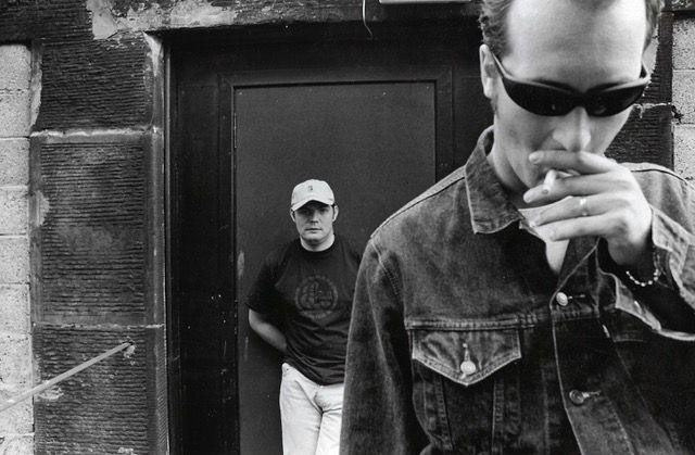 A black and white photo of two men standing in front of a brick building. One is closer to the camera holding a cigarette in his mouth and the other is behind standing in front of a door.
