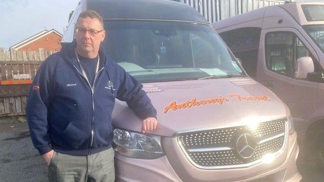 Richard Bamber, who is wearing a blue zip-up fleece and jeans, leans on the bonnet of a light purple mini-bus with the Anthony's Travel logo painted onto the bonnet in orange lettering. 