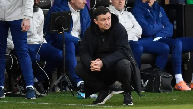 Paul Heckingbottom crouching down in the dugout wearing a long black jacket, a black top, black trousers and black and white trainers.