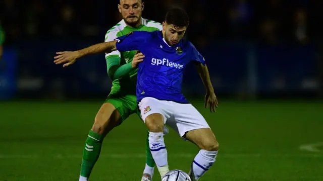 Aaron Henry shields the ball from a Yeovil Town player, wearing Wealdstone's blue kit with white shorts and socks