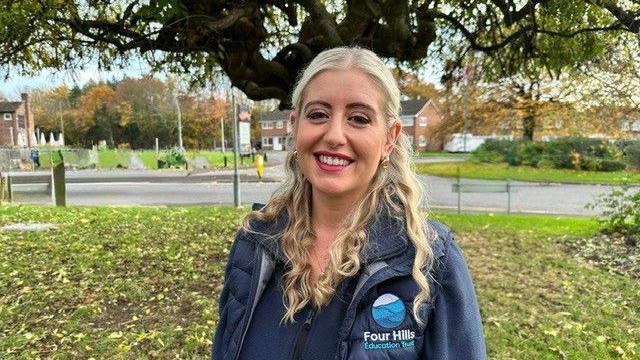 A woman with blonde hair and a blue coat standing on a grassy area with a tree behind her and leaves on the floor. Behind that there is a road and a roundabout.