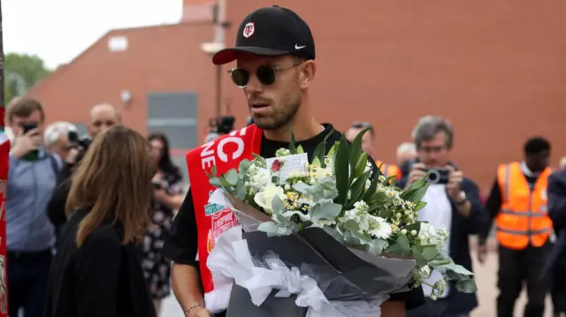 Jordan Henderson, wearing a black cap and sunglasses, carries a bunch of flowers. He also has a Liverpool scarf on his shoulder.