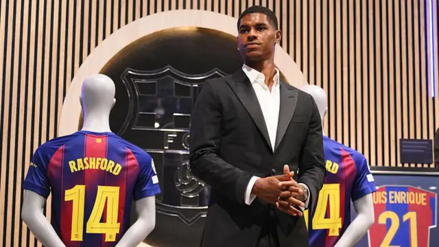 Marcus Rashford, wearing a black jacket and white shirt is unveiled at Barcelona next to a Barcelona shirt with a 14 Rashford shirt