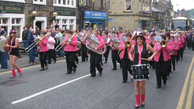 A brass band, made up entirely of female musicians wearing pink blazers and black shirts and trousers, walk up Uppermill's High Street as part of the village's annual brass band contest