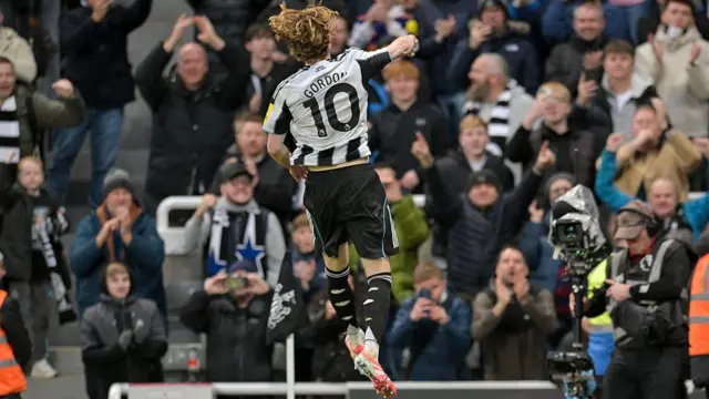 Anthony Gordon, with his back to the camera, jumping high to celebrate a goal. Newcastle fans in the crowd are out of focus in the background.
