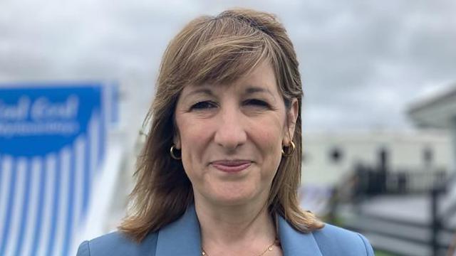 Rachel Reeves standing a smiling at the camera. She is wearing a gold necklace, gold hoop earrings and a light blue blazer.