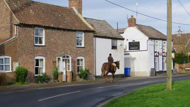 A rider on a brown horse is seen on a road in the centre of the image in front of a row of old brick and white-washed houses, including a pub, with looks to date from the 18th or 19th Centuries. To the right, a telegraph pole stands on a green grass verge.