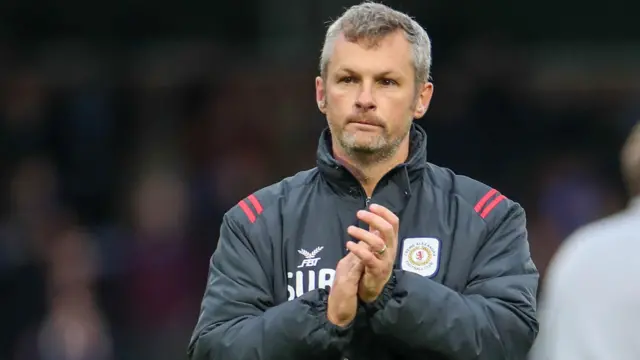 Kenny Lunt applauds the crowd after a game for Crewe. He is wearing a dark grey club coat.