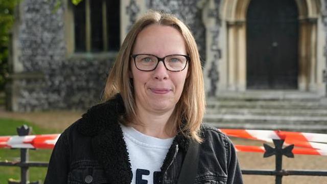 A woman smiling at the camera. She is wearing black frame glasses with a black denim jacket and a white top underneath. She is standing in front of the church.