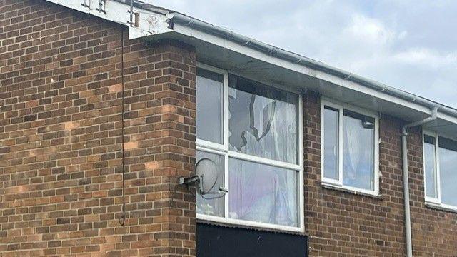 A large smashed window of an upstairs flat. The modern flat building is made of brown brick; the pane of glass in a large window at the end of the property has been badly broken.