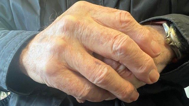 An elderly man's hands crossed, with a watch and dark jacket sleeves visible.