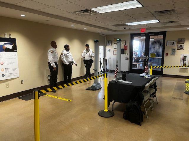 Yellow and black rope forms a line where visitors to the tabulation centre must queue to go through a metal detector. Three armed security guards stand near the detector. A black folding table sits nearby with a metal detector wand. 