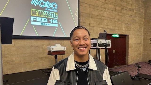 Kay Greyson smiling at the camera. She's wearing a black and white leather jacket and standing in front of a stage with a screen reading 'MOBO Awards Newcastle- Feb 18 2025.'