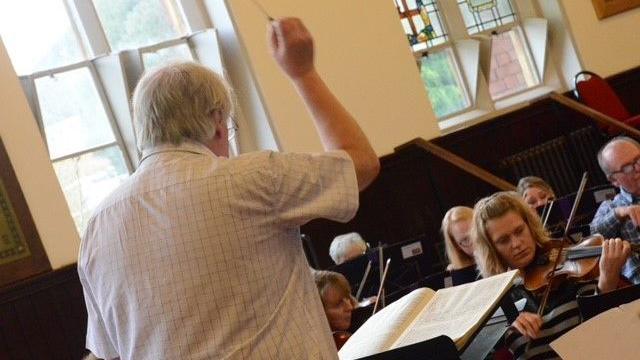 A man in a white shirt is stood at the front of a group of musicians. There is a book of sheet music in front of him, and he is hlding a stick above his head with his right hand.