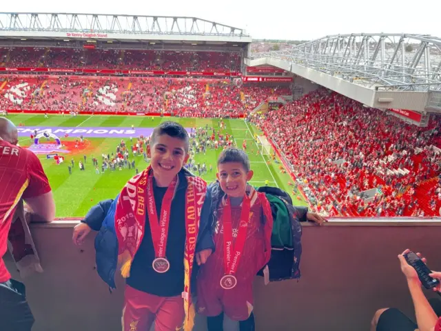 Two boys stand at Anfield with Premier League winners' medals around their necks and the pitch behind them