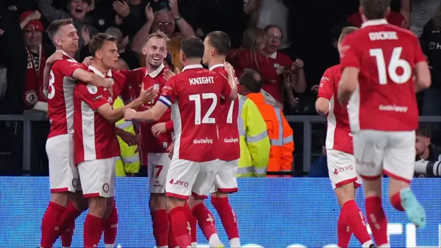 A group of seven Bristol City players wearing red t shirts, white shorts and red socks hug each other in celebration after scoring against Derby County