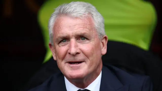 Mark Hughes in a suit looks ahead and smiles from a dugout