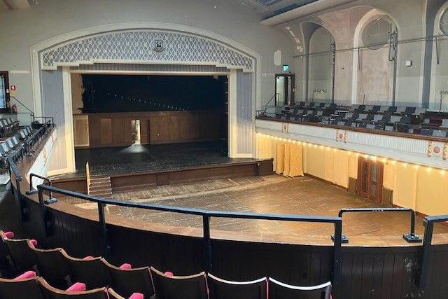 The theatre has a brown wooden floor with brown steps leading onto the stage. There is a blue surround with white pattern. Chairs are in the foreground.