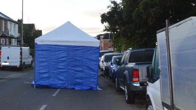 A blue and white forensic tent in the middle of a residential street. Vehicles are parked either side