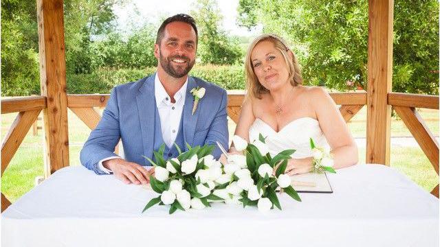 A man in a blue wedding suit with a white shirt sits next to a woman in a wedding dress at a table which has a centrepiece made of white flowers, behind them the wooden beams of an outdoor structure are visible, and further in the distance are trees and hedges