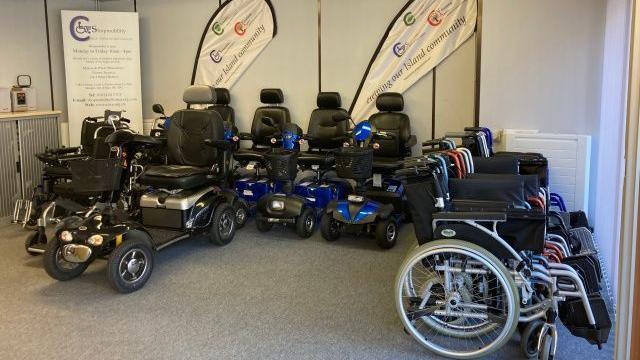 A room with mobility scooters and wheelchairs, with two flags that read Serving Our Island Community and a banner with the word Shopmobility on it in the background.