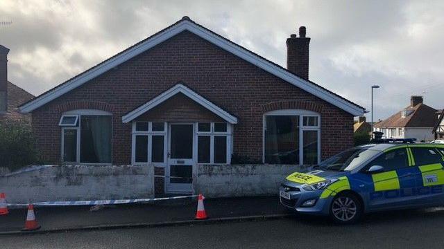 A police car parked outside a bungalow. The property has police tape and cones in front of it