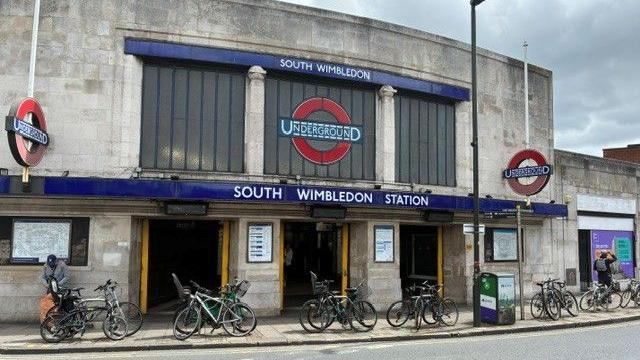 The exterior of South Wimbledon Underground station, a stone building with Underground roundels and station signage, with bicycles parked outside.