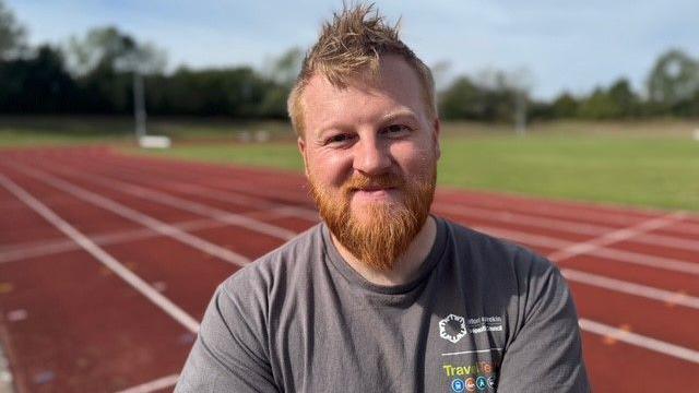 A man with ginger hair and a beard in a grey T-shirt on a running track with grass in the background