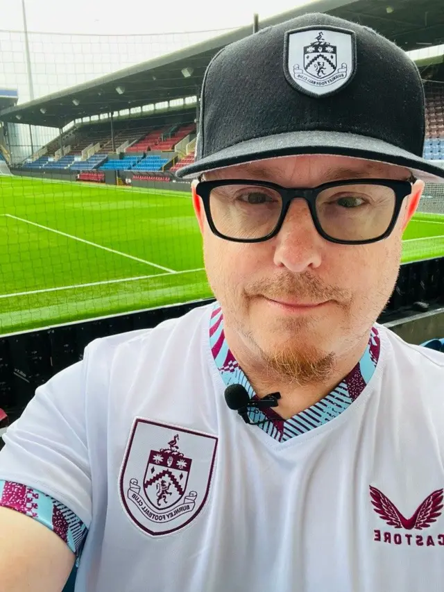 A selfie of a man with glasses and a Burnley cap, wearing a white Burnley away shirt