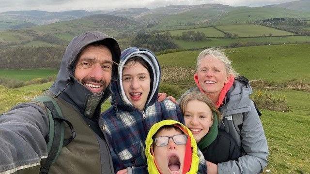 Yn y llun yma mae Rhodri Lloyd-Williams yn gwenu gyda'i wraig a'i dri o blant. Mae'r teulu tu fas, yn yr awyr agored, a gyda thir gwyrdd tu ol iddyn nhw.