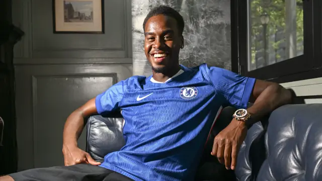 Jamie Gittens sits on a black leather sofa in a blue Chelsea shirt