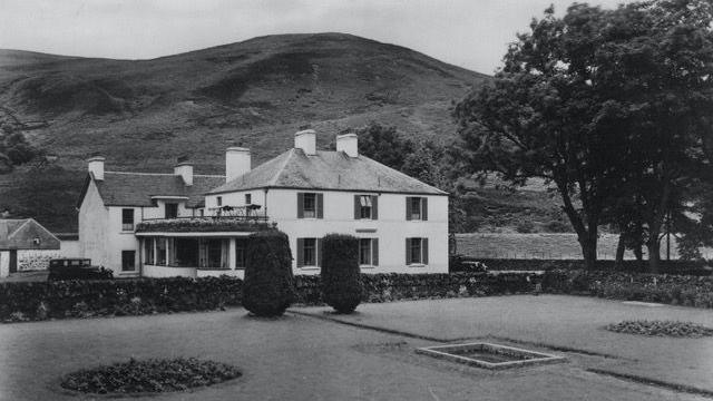 An old black and white picture of the Crook Inn in Tweedsmuir - a white building in among trees with hills in the background