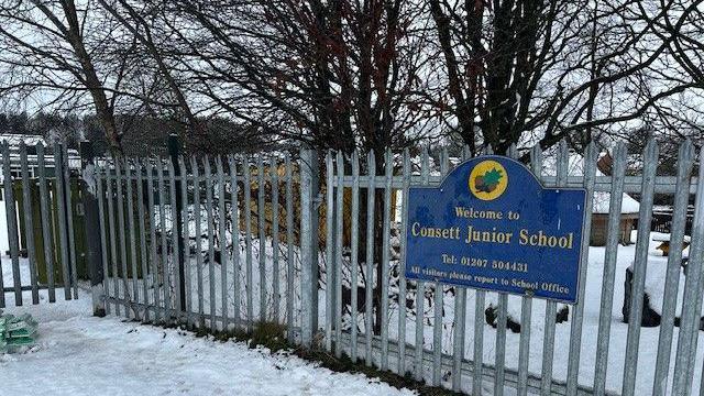 Snow-covered grounds. Metal fence with blue sign which says "Welcome to Consett Junior School". There are trees in the background. 