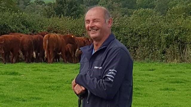 Llun o Alun Thomas yn gwenu y tu allan yn y cae. Mae o'n gafael mewn ffon a gwelir gwartheg yn y cefndir yn y cae.