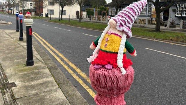 A knitted Christmas elf figurine dressed in a long pink hat sits on bollard beside a road in Darlington