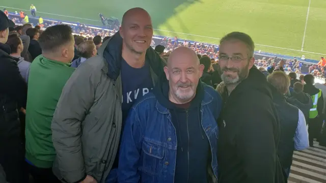An image of three men at Portman Road with sun on the pitch.
Two of the men are bald wearing a green jacket and a denim jacket. The other man has grey hair, a grey beard and is wearing a black hooded top.