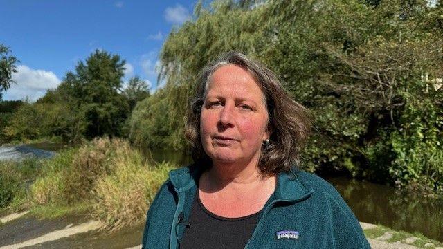 Alison Caffyn standing by the River Teme in Ludlow. She has brown shoulder-length hair and is wearing a black t-shirt and dark green cardigan. Behind her is the river and green trees and foliage. 