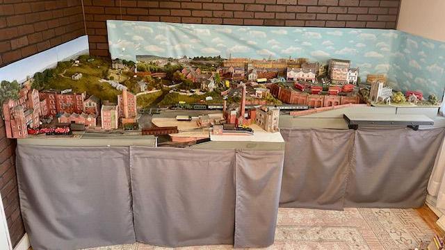 A view of the whole model which includes miniature red brick buildings, cars and a train seen running through it. There are hills rolling up to the left of the scene and churches are also seen while in the background there are clouds painted on a sky and the wall of the room behind the whole thing.