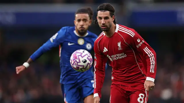 Dominik Szoboszlai, wearing Liverpool's red home shirt and shorts, concentrates on a bouncing ball, as Chelsea's Malo Gusto runs towards him in the background