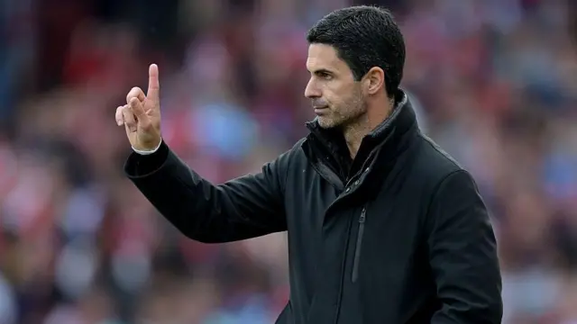 Mikel Arteta wearing a black coat with his arm outstretched and his finger pointing upwards