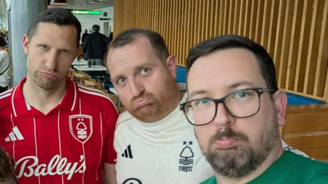 Three people are stood next to each other looking towards the camera with a sad face. They are all wearing different colour football shirts and are standing in front of a wooden background.
