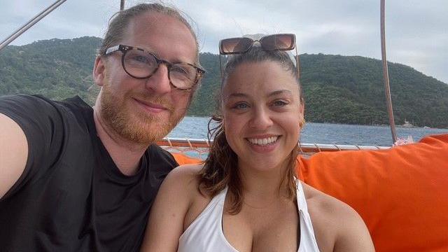 A man with a beard and long hair pulled back, wearing glasses, is sat on the left wearing a black t-shirt. Next to him, on the right of the frame, is a smiling woman. Their is an ocean and green island behind them. Both are sat on a boat.