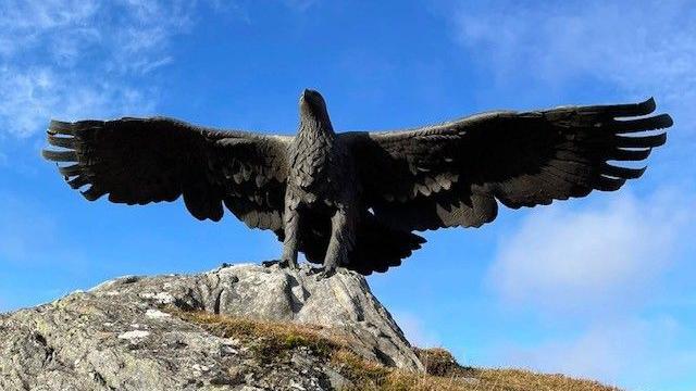 A giant eagle statue on a rock. The eagle is a dark colour and is standing on a light coloured rock. Small bits of greenery can be seen to the side. It is in front of blue sky and cloud.