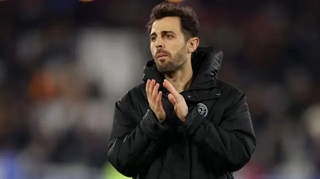 Bernardo Silva, wearing a black substitutes' jacket and applauding towards Manchester City fans