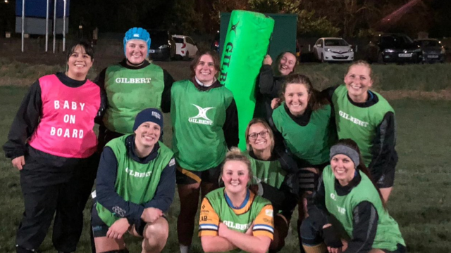 A group of women rugby players all in green vests except Trish, whose is pink and says 'Baby on Board' on it