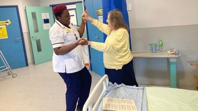 A woman in white and blue nurse's uniform is holding hands with a patient, who is in a yellow jumper and dark blue skirt. They are dancing in a hospital room, and are smiling.