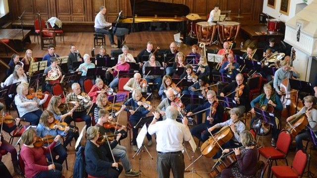 A man in a white shirt is stood in front of a group of about 40 musicians with instruments. He is holding a conductor's stick in his right hand, and is holding his left hand up to indicate instructions to the players.