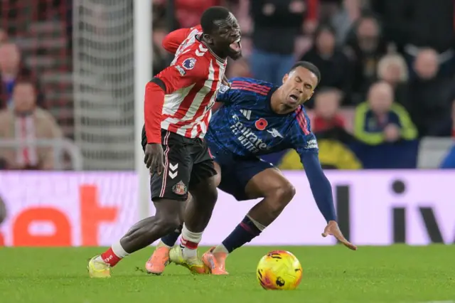 Brian Brobbey of Sunderland going to ground in pain under a challenge from Arsenal's Gabriel, who is standing on his foot with the ball just away from them in the foreground. The crowd is out of focus in the background.