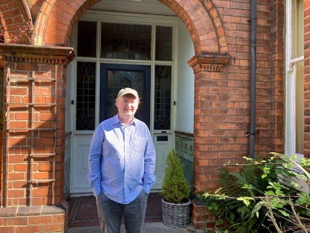 Stephen Ingram is smiling. He wears a blue shirt, grey trousers and grey cap and is standing in front of the door of a red-brick house